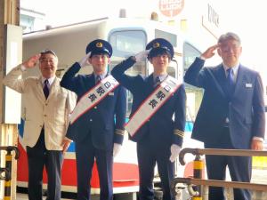 駅員の制服を着た藤井聡太叡王と小山怜央四段が「一日駅長」と書かれた襷を掛け敬礼し、その二人の両端にも敬礼している男性が立ち4人が並んでいる記念写真