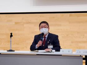 協議会会場で市長が資料が置かれたテーブルの前に座りマイクを片手に持ちながら意見を述べている写真