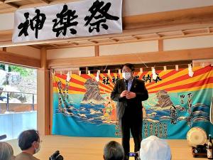 入口上部に「神楽祭り」と書かれた紙が貼られた神楽堂の中に立ちマイクを持って話をする様子の市長の写真