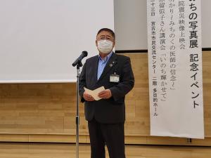 イベントの題名が書かれた垂れ幕が掛けられ、スクリーンが設置された会場でスタンドマイクを前に話をする市長の写真