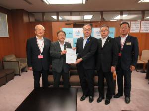 バックパネルや応接セットが設置された室内で、国土交通省の吉岡幹夫技監と市長が要望書を一緒に持ち、3名の関係者が横一列に並んでいる写真