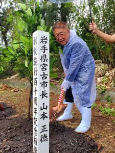 雨が降るなかスタッフに傘をさしてもらいレインコートを着た市長が「村制施行110周年記念」と書かれた標柱の傍で植樹を行っている様子の写真