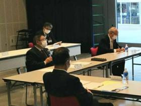 会議の会場で市長が自席に座り、マイクを片手に持ちながら話をしており他の参加者は資料を見ながらきいている写真