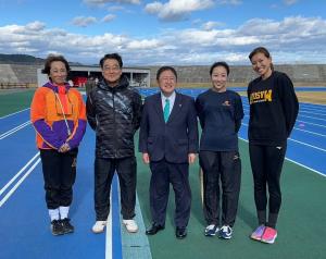 陸上競技場でスーツ姿の市長を中央に、大村先生や3名の女性講師の方が横一列に並んでいる記念写真