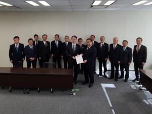 茶色の長机が設置された室内で、文部科学省研究振興局長の森晃憲氏と市長会の代表が一緒に要望書を持って前列に立ち、後列に市長を含む12名の関係者が横一列に並んでいる写真