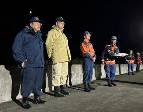 太陽が沈み真っ暗な中堤防に市長や2名の関係者が横一列に並び、市長が挨拶を行っている様子の写真