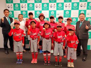 宮古市のバックボードの前で赤いユニフォームを着たサンライズ千徳スポーツ少年団の皆さんが2列に並びその両端に市長と副市長が立ち一緒にガッツポーズをし記念撮影をしている写真