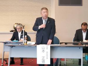「山本正徳」と書かれた名前札が垂らされた机と椅子の間に立ち、マイクを持って話す様子の市長と、後方で席に着き手元を見る男性2名の写真