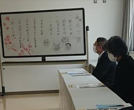 ホワイトボードにイラストや手書きで式次が書かれた入学式の会場で、一番前の席に座っている市長がホワイトボードを見つめている写真