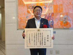 ロビーの壁に飾られたオレンジ色の作品をバックに「国土交通大臣表彰」を受賞した市長が、表彰状を両手で持ち立っている写真