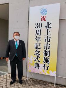 屋外に設置された「北上市市制施行30周年記念式典」と書かれた看板の左側に立つ市長の写真