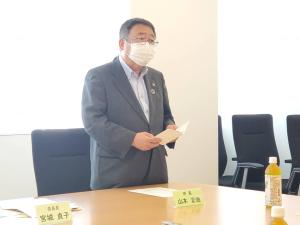 ネームプレートが置かれた机と椅子の間に立ち、両手で資料を持ち話をしている様子の市長の写真