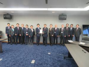 青色の絨毯に長机が設置された室内で、経済産業省副大臣の中谷真一氏と市長会の代表が一緒に要望書を持って中央に立ち、市長を含む12名の関係者が横一列に並んでいる写真