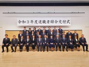 「令和3年度退職者辞令交付式」と書かれた横断幕が舞台後方上部に掲げられ、舞台上には1列目に椅子に座った13名が並び、2列目には10名が立って並び、3列目にも8名が立って並んでいる記念写真