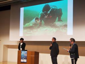 ダイビング中の写真が映し出されたスクリーンの前でマイクを持ち話している様子の3人の男性の写真