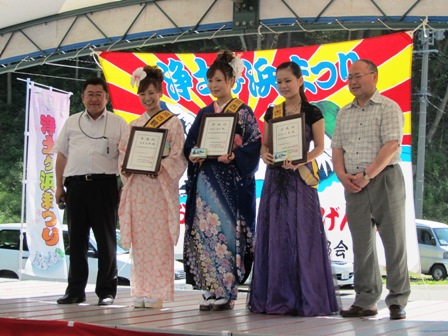 特設ステージの上で手に賞状を持った着物やドレスを着た3名の女性と2名の男性が記念撮影している写真
