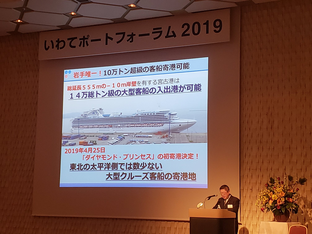 「いわてポートフォーラム2019」と書かれた横断幕の下に資料が映し出さされたスクリーンが設置された会場で、右側の檀上に立つ市長が話をしている写真