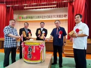 「多良間村制施行110周年記念式典並びに祝賀会」と書かれた横断幕が飾られたステージ下で、玉城沖縄県知事と伊良皆光夫村長や市長と2名の関係者がそれぞれ木槌を持ち樽酒を囲んでいる鏡開きの様子の写真