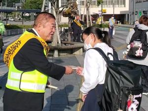 反射材のベストを着て、その上から交通安全の襷をかけた市長が、リュックを背負った女子生徒に交通安全街頭啓発運動をしている写真