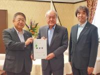 白いテーブルクロスがかけられた机の前で、市長を含む男性3名が並んで立ち、市長と男性で要望書を持っている写真