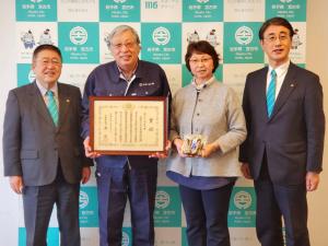 バックパネルの前で、市長と副市長の間に、丸友しまか有限会社の男性が額縁に入った賞状を持ち、女性が受賞した商品を持って並んでいる写真