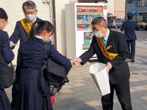 黄色のたすきを肩に掛けた市長と男性が、通学中の女子生徒に啓発グッズを手渡している様子の写真