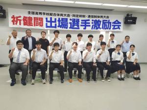 「祈健闘 出場選手激励会」と書かれた横断幕が壁の上部に吊るされた部屋で、前列に制服を着た男女の学生が8名座り、後列に学生6名と市長を含む関係者の方々4名が立って並び全員が片腕を曲げ握った拳を顔の横に置いてポーズを決めている写真
