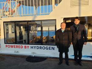 POWERED BY HYDROGENと書かれた停泊中の水素燃料旅客船の前で市長と男性が並んでいる写真