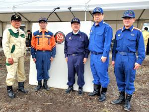 テントと2本のマイクが設置された演台の前で、作業着を着用した市長を中央に4名の関係者が横一列に並んでいる写真