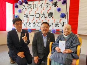 右手の人差し指を上に掲げポーズをとる関係者の男性と市長が中腰になり、市内最高齢者である久保田イチさんが目録を持って椅子に座っている様子を写した写真