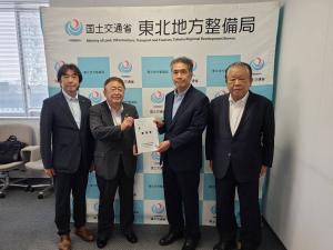 「国土交通省 東北地方整備局」と書かれたバックパネルの前で要望書正面に向け一緒に持つ市長とスーツを着た男性、その2人の両端に立つ関係者の男性2名の記念写真