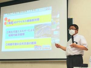 画面が映し出されたスクリーンを指さしながら、左手にマイクを持ち話をしている様子の市長の写真