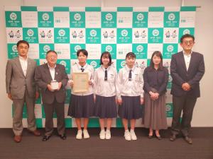 バックパネルの前で制服を着た女子中学生3人を中心に、市長と関係者3人が横一列に並ぶ記念写真