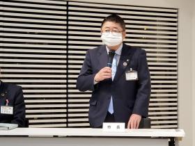 「市長」と書かれたプレートが置かれたテーブルの前に立ち、マイクを片手で持った市長が市民へ説明している写真