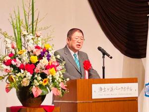 演台横に赤、黄色、白、オレンジ、紫の色とりどりの花が活けられた花瓶が飾られ、赤色の胸章リボンをつけた市長が演台のマイクを使いながら挨拶をしている写真