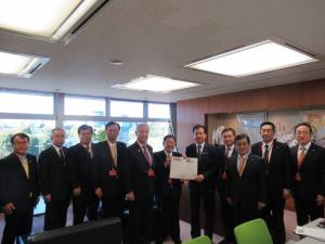 大きな窓ガラスに茶色の重厚な壁のある部屋で、国土交通大臣政務官の小鑓隆史氏と市長が一緒に要望書を持って中央に立ち、9名の関係者が横一列に並んでいる写真