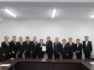 茶色の長机が設置された会議室の壁側に農政局農産政策部長の松本平氏と市長が一緒に要望書を持って中央に立ち、10名の関係者が横一列に並んでいる写真