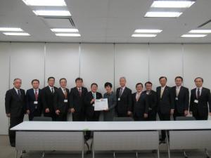 白色の長机が設置された会議室の壁側に研究振興局長の塩見みづ枝氏と市長が一緒に要望書を持って中央に立ち、11名の関係者が横一列に並んでいる写真