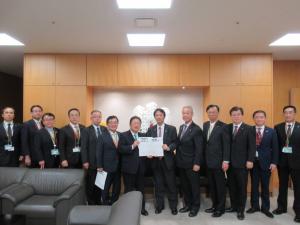 黒色の革張りのソファが設置された室内で内閣府副大臣の工藤彰三氏と市長が一緒に要望書を持って中央に立ち、11名の関係者が横一列に並んでいる写真