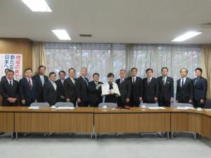 レースのカーテンがひかれた会議室の窓際に幹事長代理の稲田朋美氏と市長が一緒に要望書を持って中央に立ち、13名の関係者が横一列に並んでいる写真