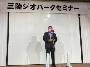 「三陸ジオパークセミナー」と書かれた横断幕が上部に掲げられた会場で、紅白リボンのついたスタンドマイクの前で話をする様子の市長の写真