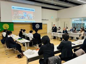 コの字がたに設置された長机に児童、生徒や関係者の方々が座り、正面のスクリーンに画像が写され、市長が起立をして挨拶をしている沖縄県多良間村派遣団報告会・解団式の写真