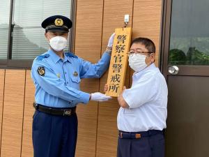 施設の入り口に「警察官警戒所」と書かれた木製のプレートを一緒に掛けている様子の制服を着た警察官と市長の写真