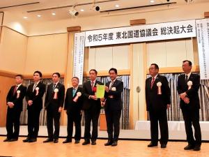 大きなホールに胸章を付けたスーツ姿の男性が8人並び中央の男性2人が要望書を持っている写真