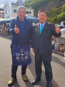 腰エプロンを身に着けた男性と市長が腕を胸の辺りまであげて手を握りしめ親指だけを上に向けポーズをとっている写真
