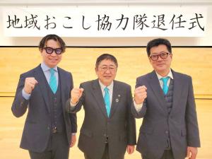 地域おこし協力隊退任式と書かれた横断幕の下で退任される吉浜知輝さんと福原隆泰さんの間に市長が入り、それぞれ右手でガッツポーズをしている写真
