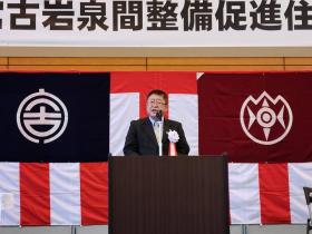 横断幕の下に 張ってある紅白の幕に岩泉町、宮古市のそれぞれの旗が張ってあるステージ上で 市長が白のリボン胸章をつけ、式辞を述べている写真