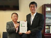 壁にテレビがかけられた部屋で、市長と男性で要望書を持っている写真