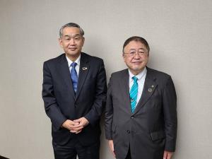 薄いグレー色の壁の前で、日本国土開発株式会社の朝倉健夫代表取締役社長と市長が並んでいる写真