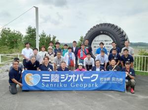 大きなタイヤのオブジェの前で、前列に並ぶ方たちがしゃがんで「三陸ジオパーク」と書かれた横断幕を広げ、腰を屈めて膝に手を置く方たちが2列目に並び、3列目には立って正面を見る方たちが横並びになって写る記念写真